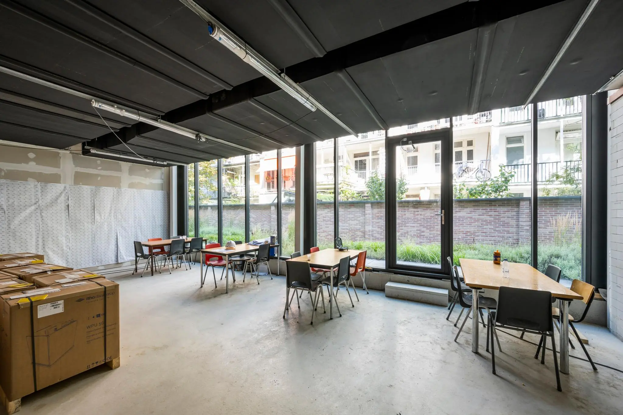 Gemeenschappelijke ruimte aan de Karel du Jardinstraat met tafels, stoelen en grote ramen met uitzicht op een binnentuin.